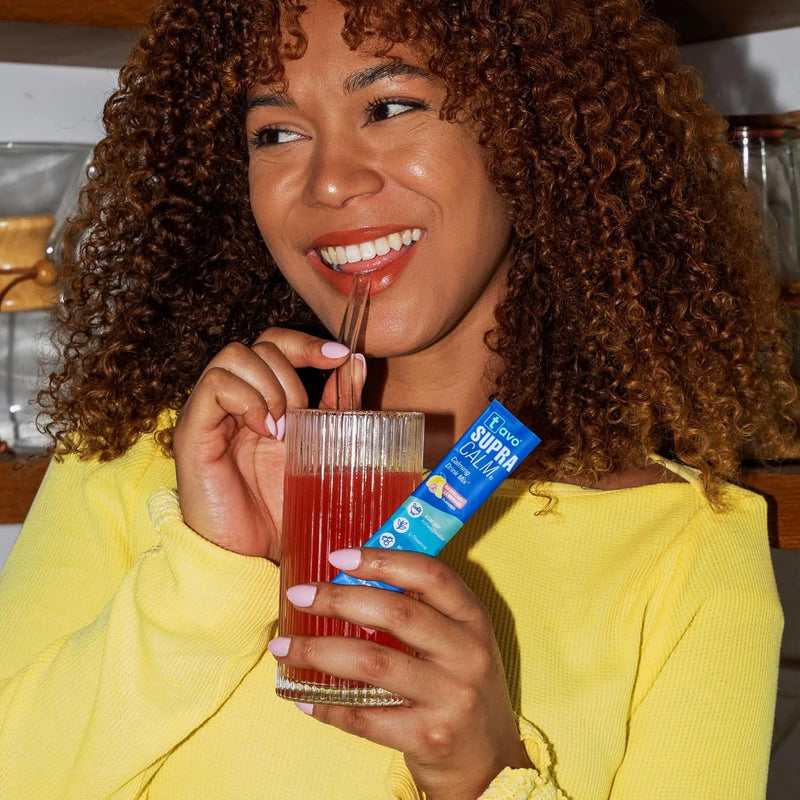 Smiling woman in a yellow top sipping a red Tavo LiquidCalm liquid calm drink and holding a stick pack in her kitchen.