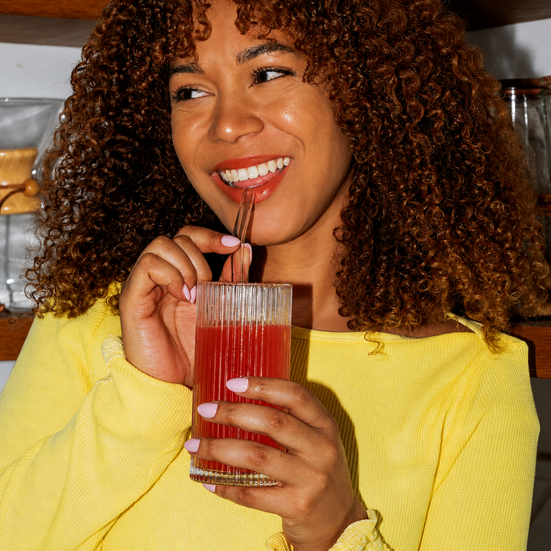 Smiling woman in a yellow top sipping a red Tavo LiquidCalm liquid calm drink and holding a stick pack in her kitchen.