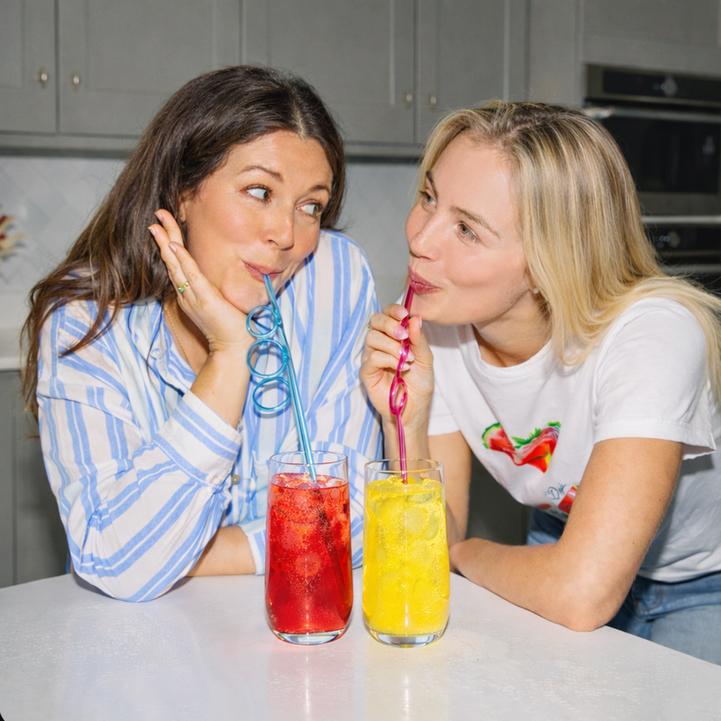 Two friends in a kitchen sipping red and yellow Tavo LiquidHydrate liquid hydrate drinks through curly straws, making daily hydration feel fun.